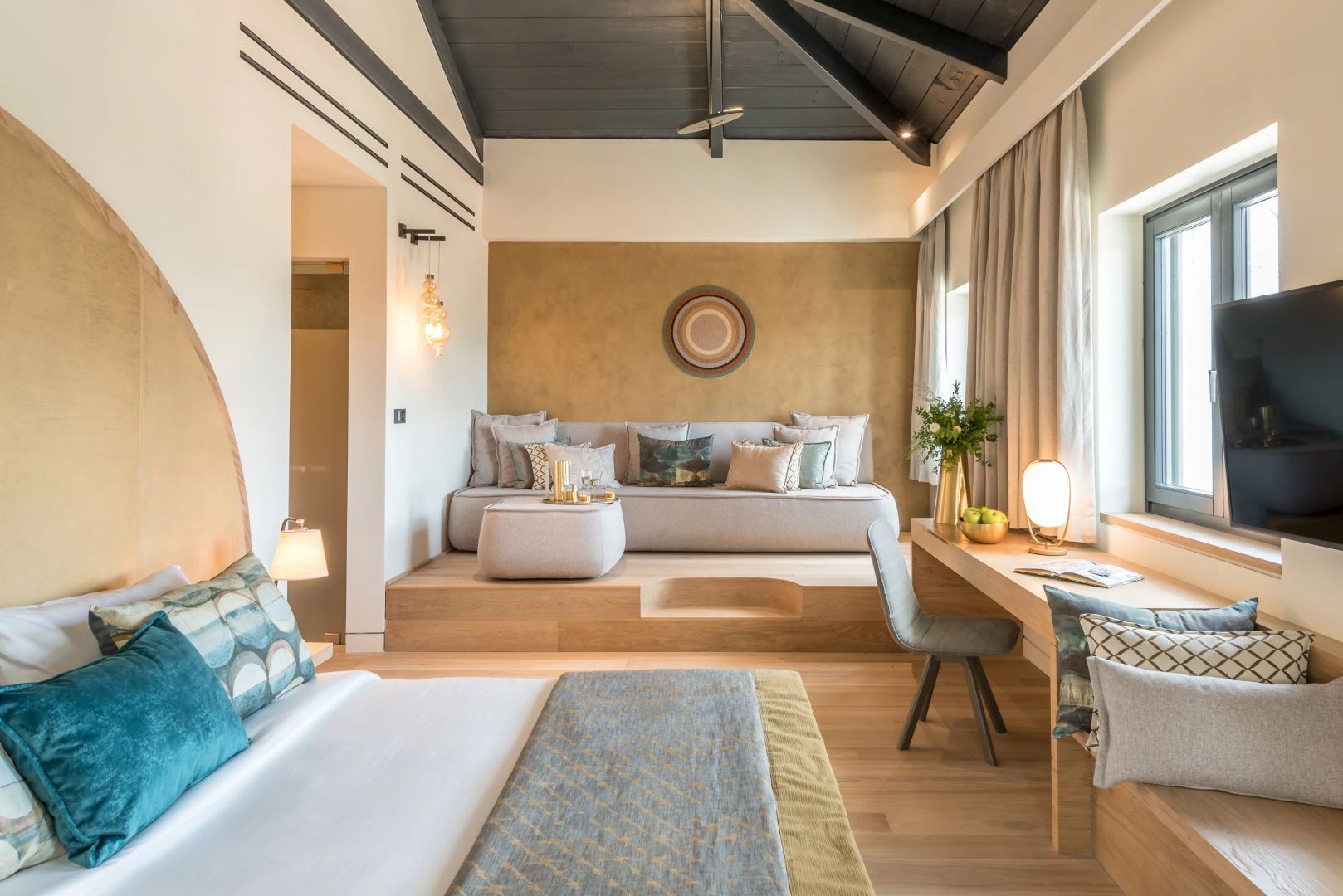 Modernes Hotelzimmer mit Doppelbett, Schreibtisch und erhöhtem Sitzbereich unter Holzgebälk.