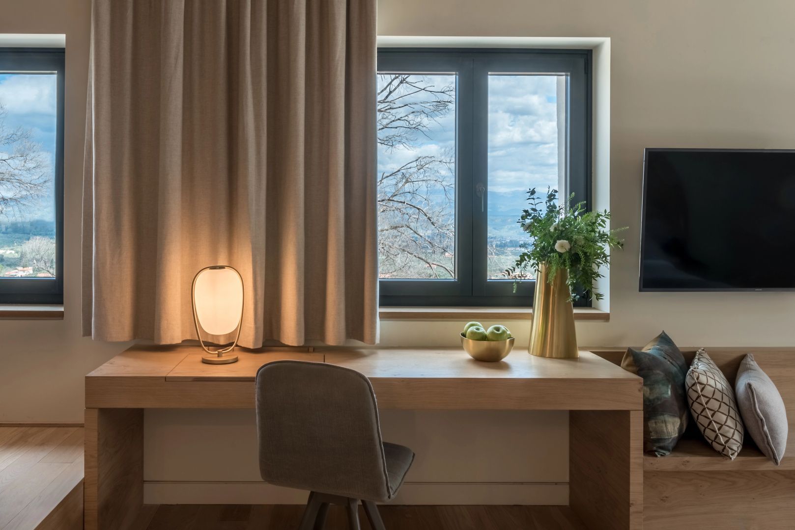 Schreibtisch im Zimmer mit Tischlampe, Blume in goldener Vase und Fensterblick ins Freie.