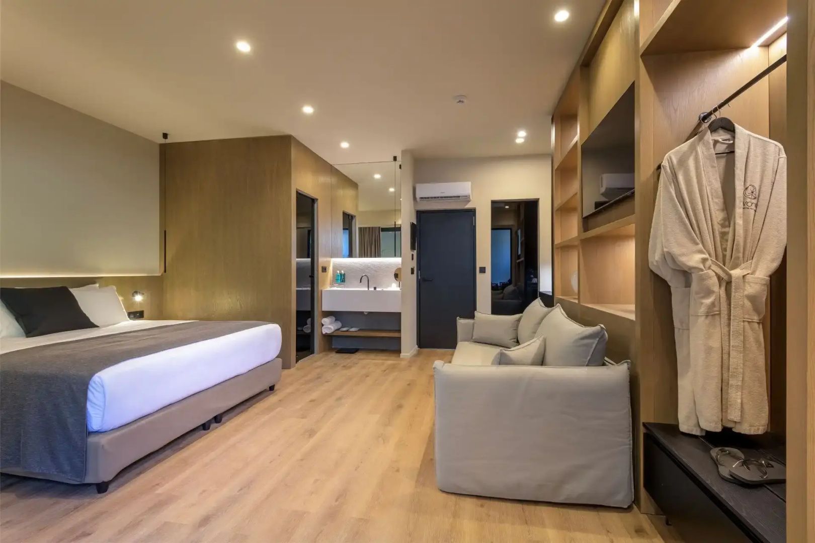Blick durch ein modernes Hotelzimmer mit hellen Möbeln, Sofa und offenem Waschbereich.