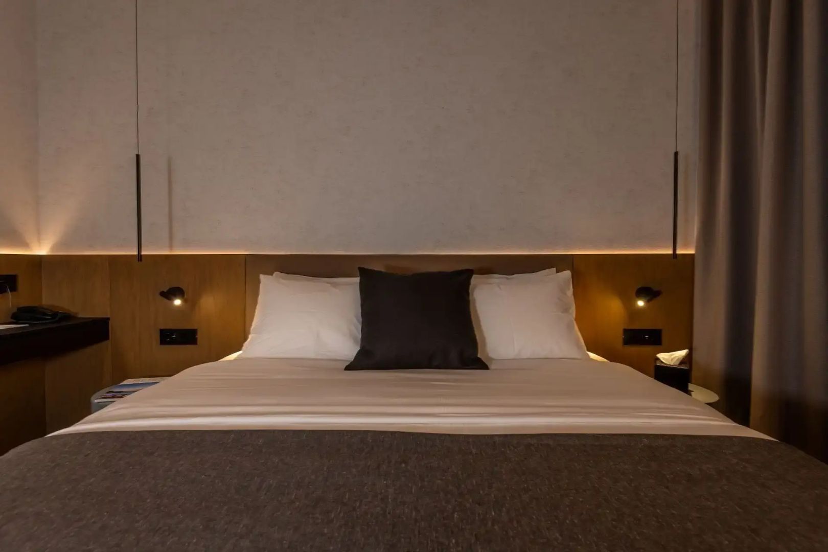 Doppelbett mit weißen Kissen und einem dunklen Kissen unter dezenter Wandbeleuchtung.