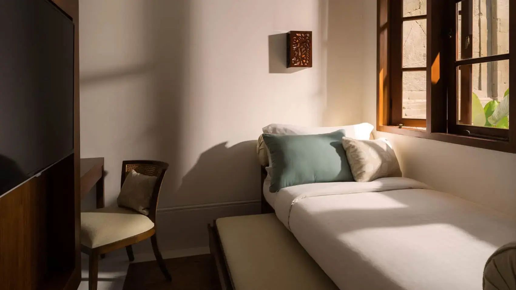 Liege mit bunten Kissen und ein Holzstuhl in einem sonnendurchfluteten Zimmer.