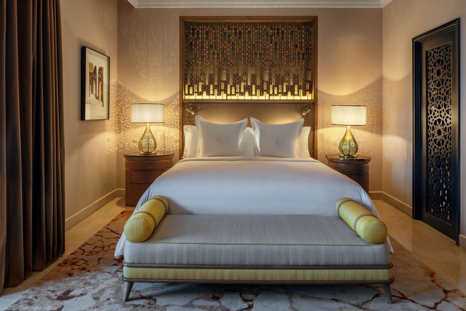 Doppelbett mit weißer Bettwäsche, kunstvollem Kopfteil und beleuchteten Nachttischen im Hotelzimmer.