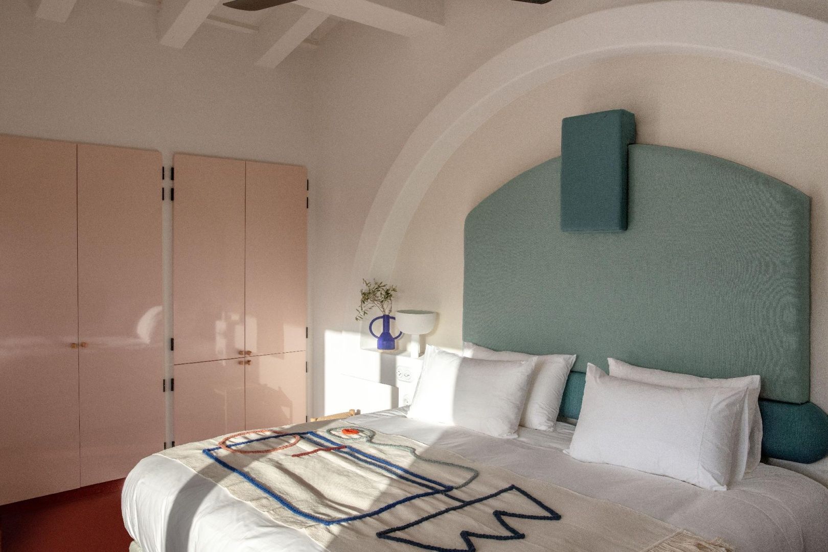 Helles Hotelzimmer mit Doppelbett, blauem Kopfteil und einem rosa Schrank.