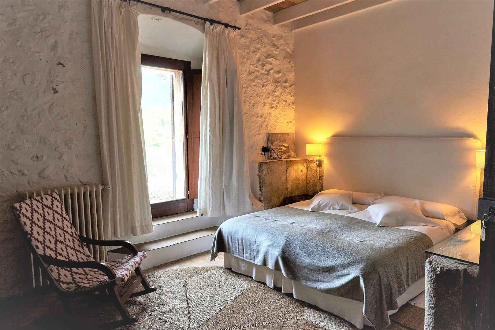 Hotelzimmer mit Doppelbett, Natursteinwand, Vorhängen und einer Tür nach draußen.