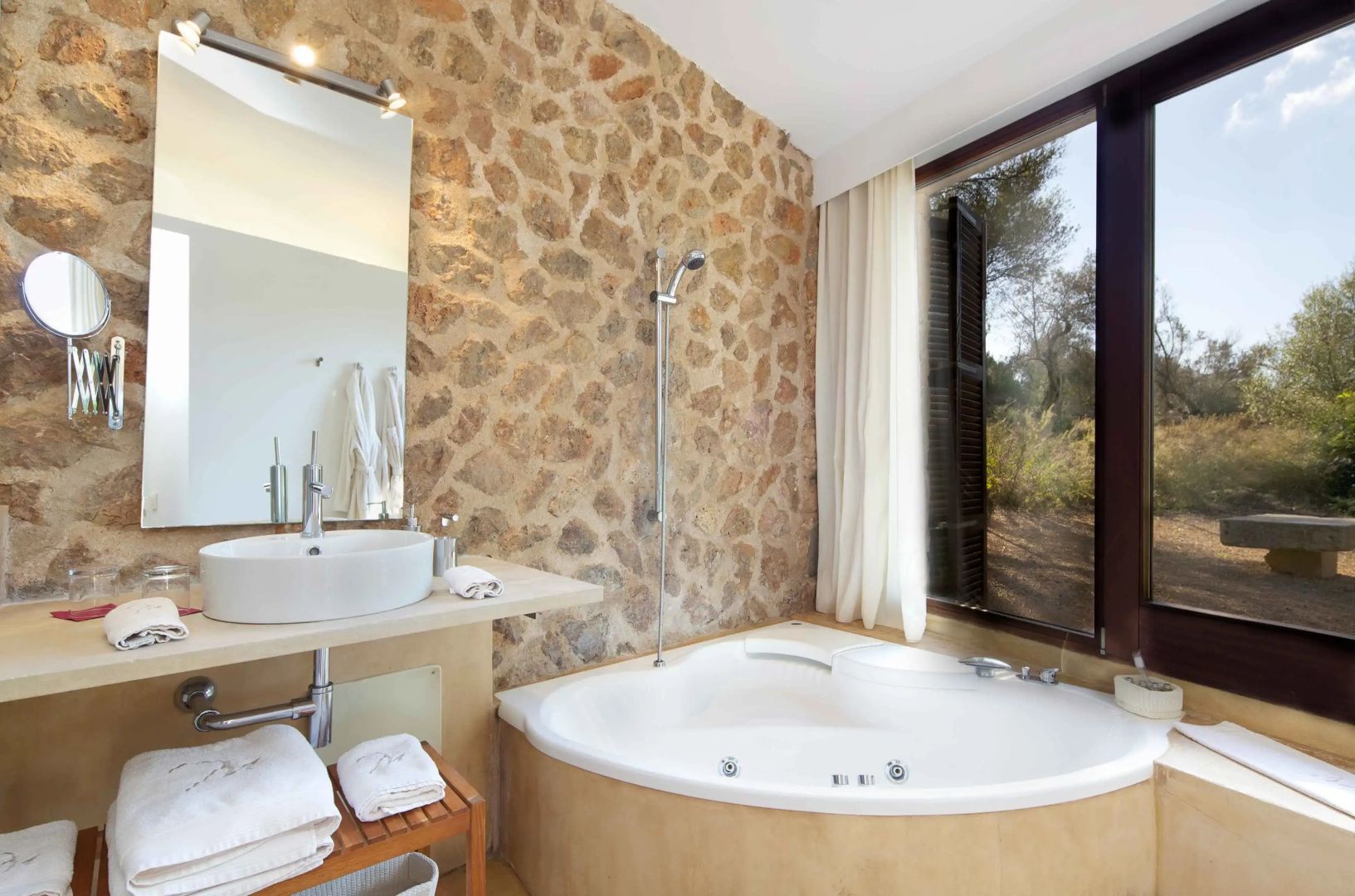 Badezimmer mit großer Eckbadewanne, Waschtisch, Spiegel und einer Natursteinwand.