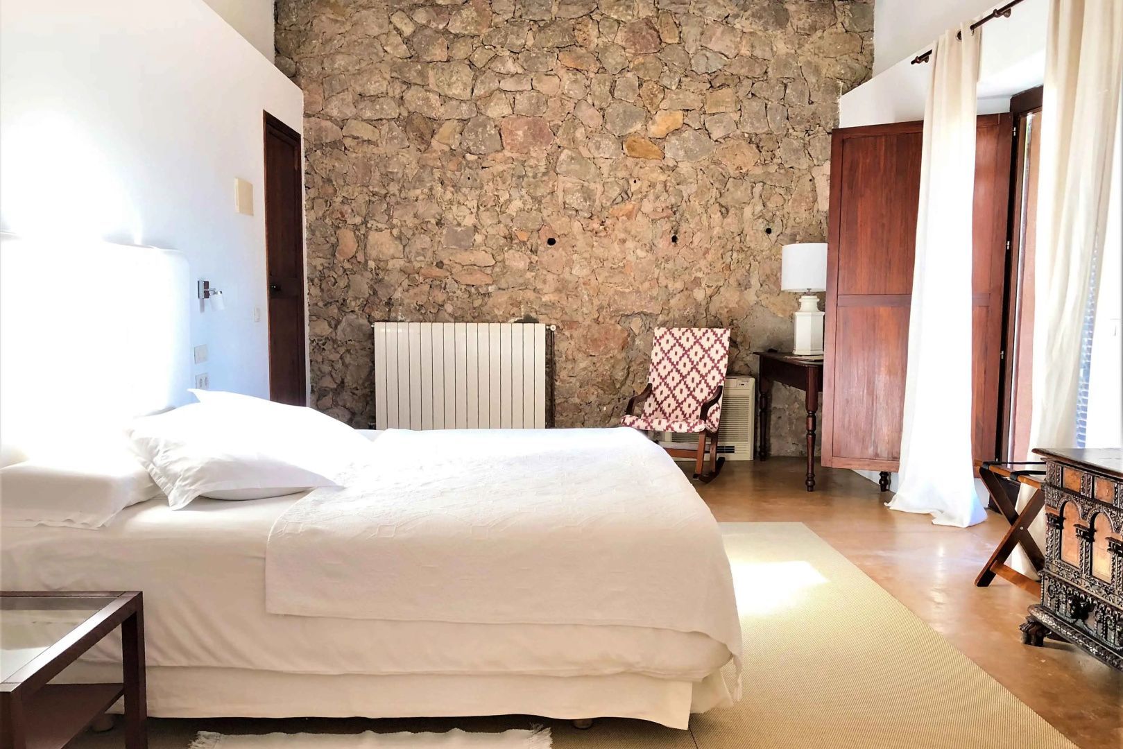 Schlafbereich mit weiß bezogenem Bett, Schaukelstuhl und sichtbarem Natursteinmauerwerk.
