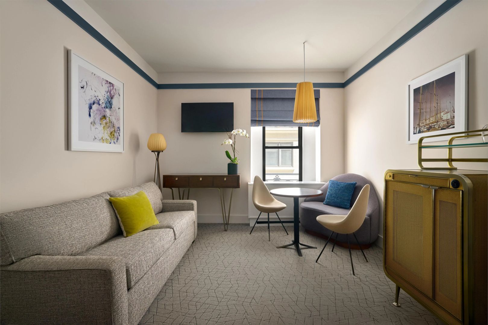 Wohnbereich eines Hotelzimmers mit grauem Sofa, Tisch, Stühlen und Fernseher an der Wand.