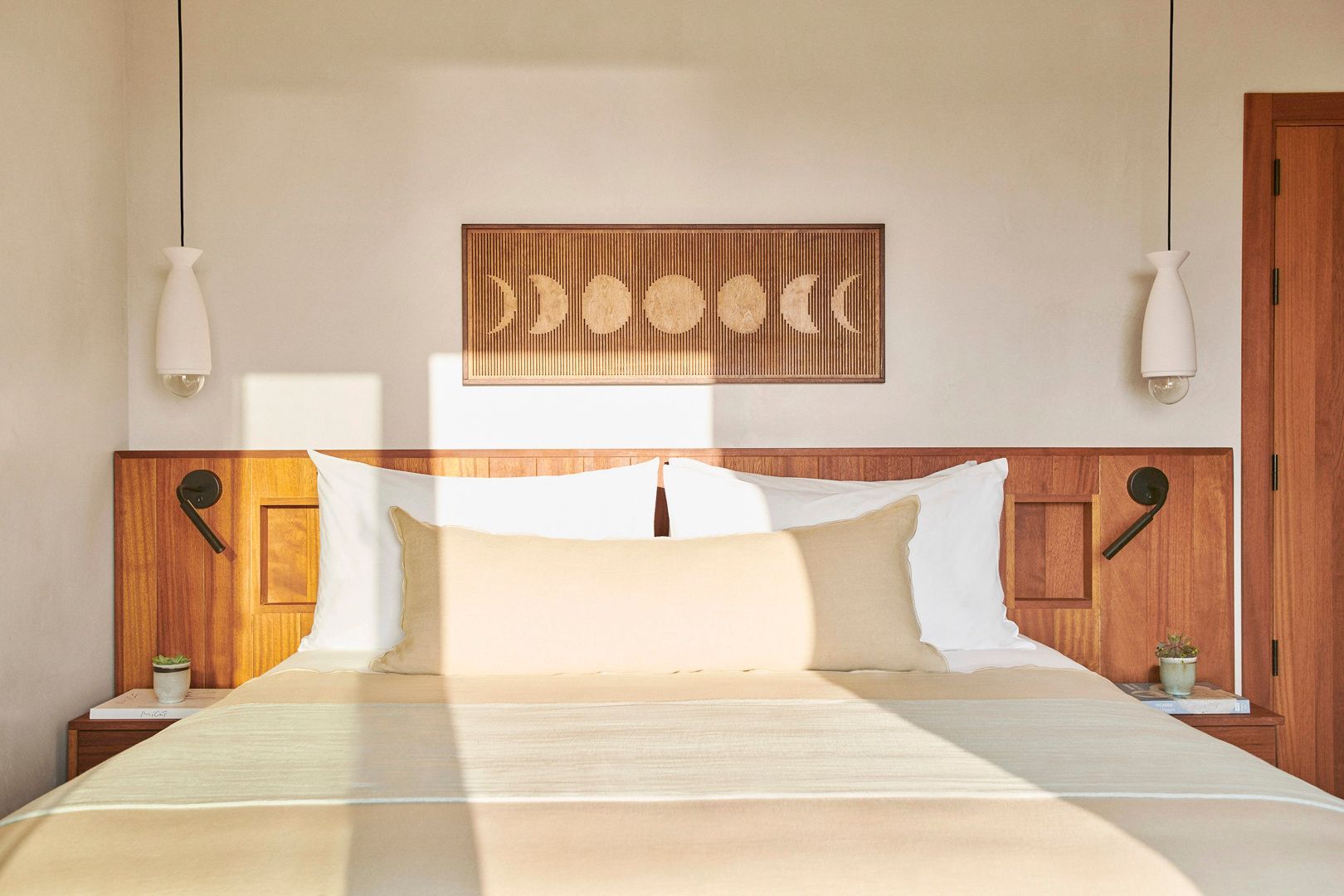 Doppelbett mit hellen Kissen, Holzkopfteil und Wanddekoration mit Mondphasen im Sonnenlicht.