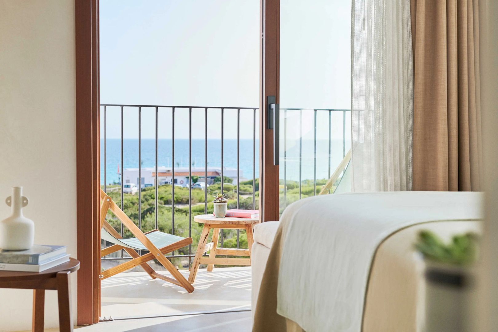 Blick vom Zimmer auf einen Balkon mit Holzstuhl und das dahinterliegende blaue Meer.