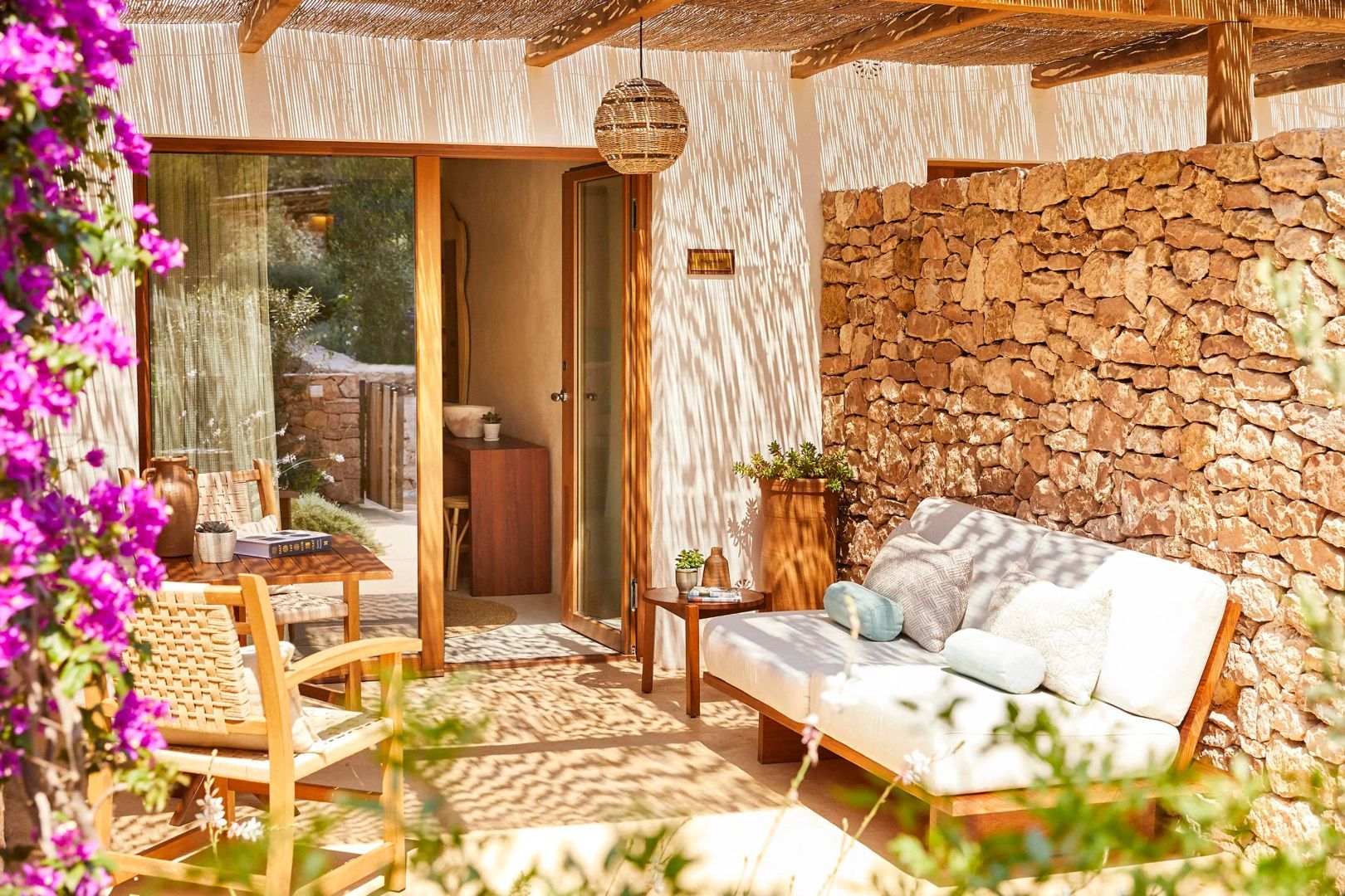 Überdachte Terrasse mit hellem Sofa und Holzstühlen neben einer Natursteinmauer.