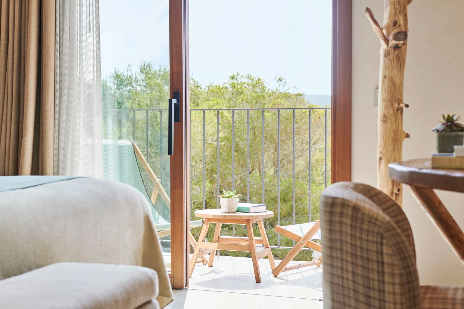 Blick vom Zimmer auf den Balkon mit Holztisch, Buch und grünem Gebüsch.