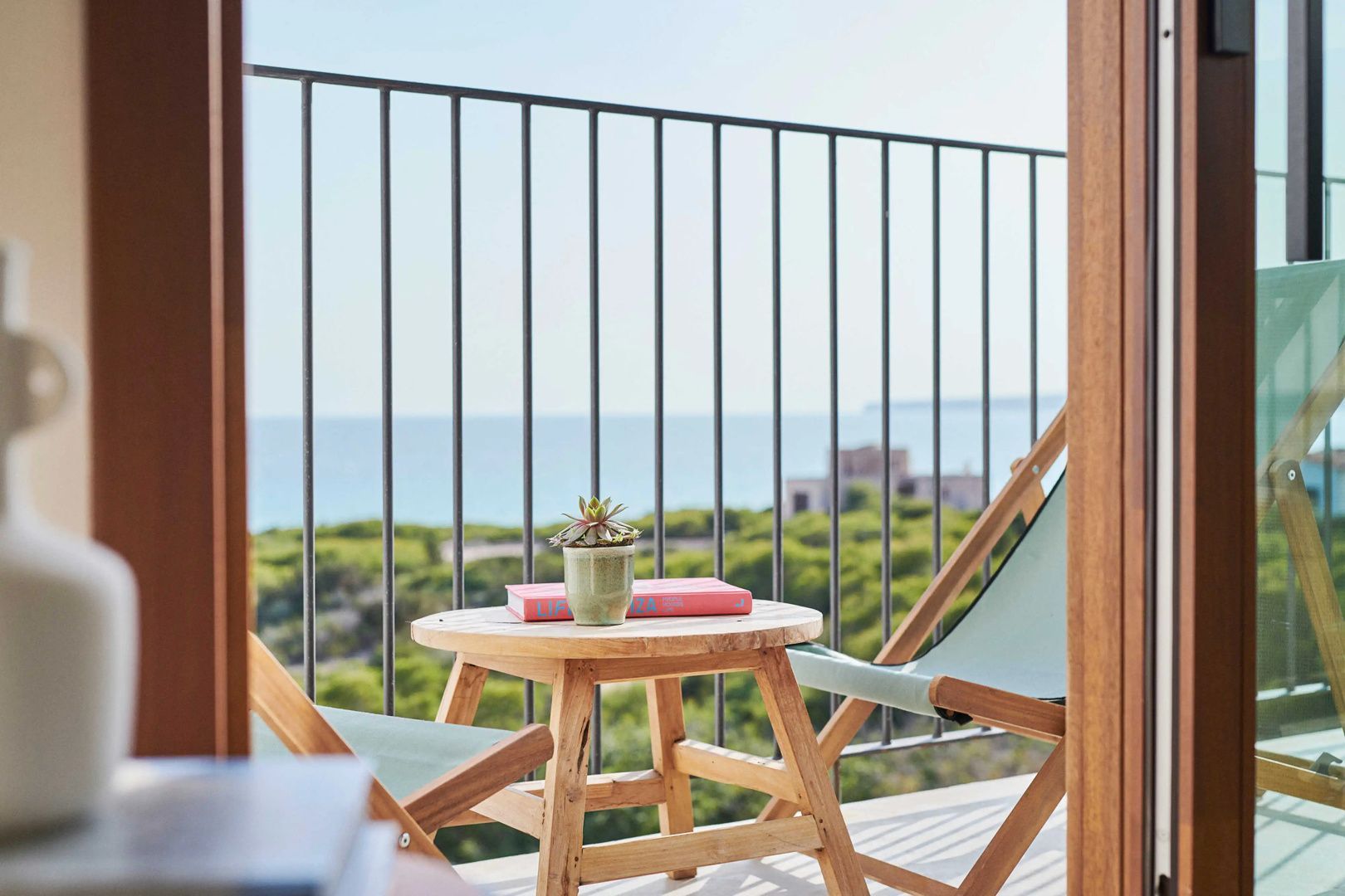 Kleiner Holztisch mit Buch und Sukkulente sowie Klappstühlen auf einem Balkon.