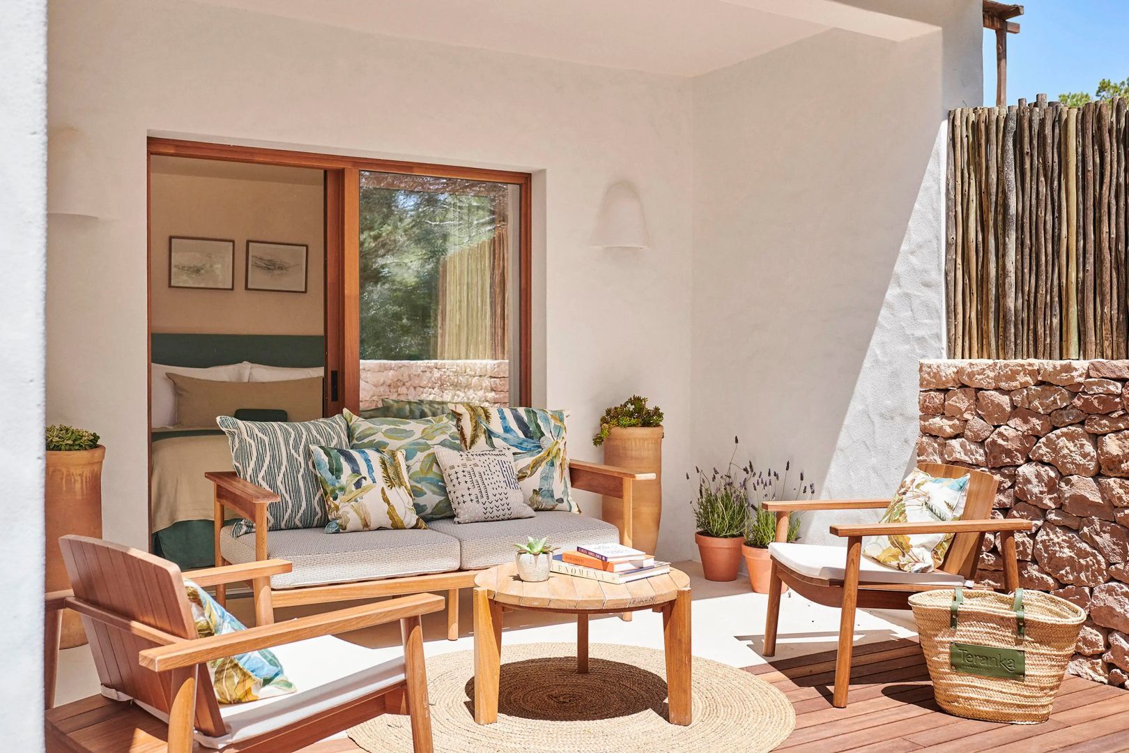 Überdachte Terrasse mit Holzmöbeln, bunt gemusterten Kissen und Topfpflanzen vor weißer Wand.