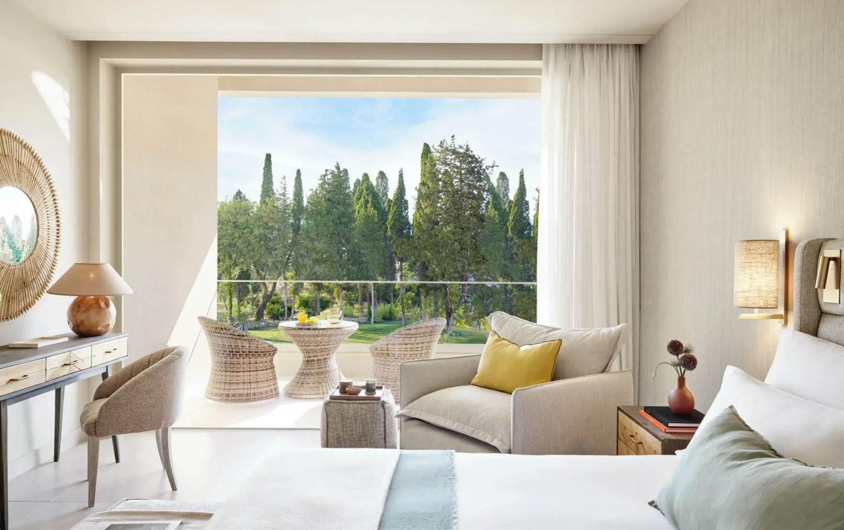 Helles Hotelzimmer mit Sessel und Blick auf eine Terrasse im Grün.