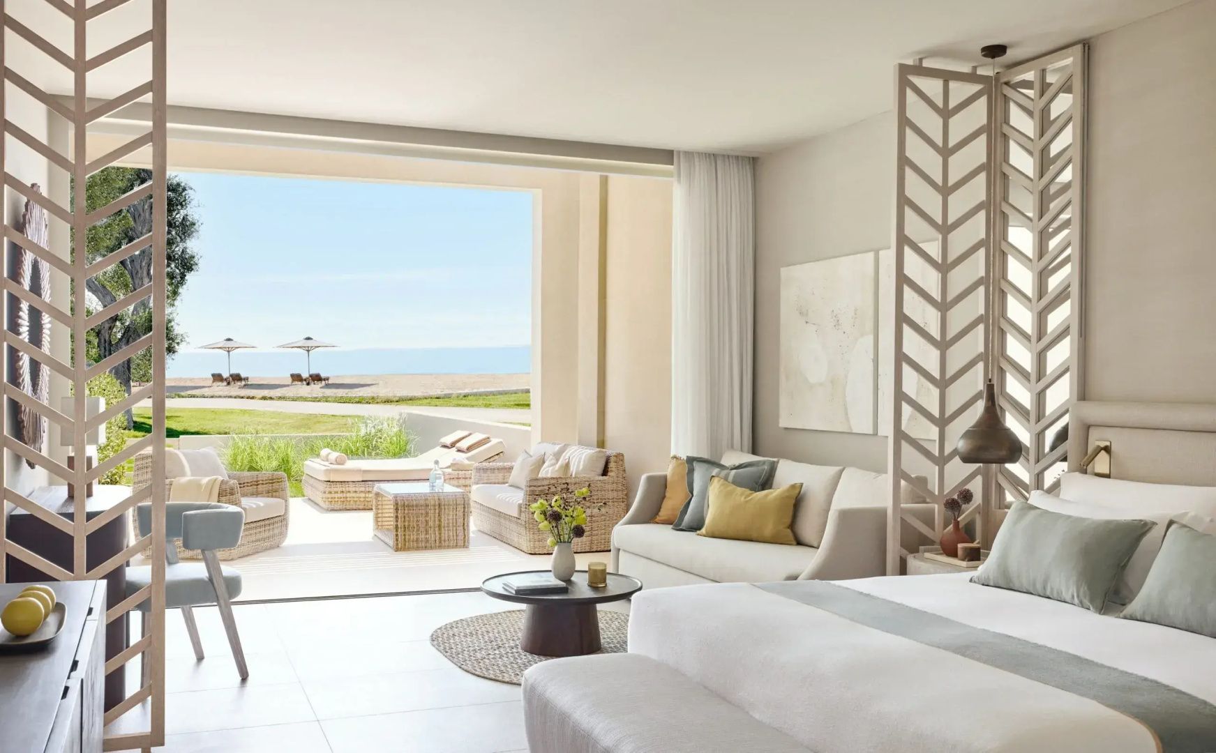 Helles Hotelzimmer mit Sofa, Beistelltisch und freiem Blick auf die möblierte Terrasse und das Meer.
