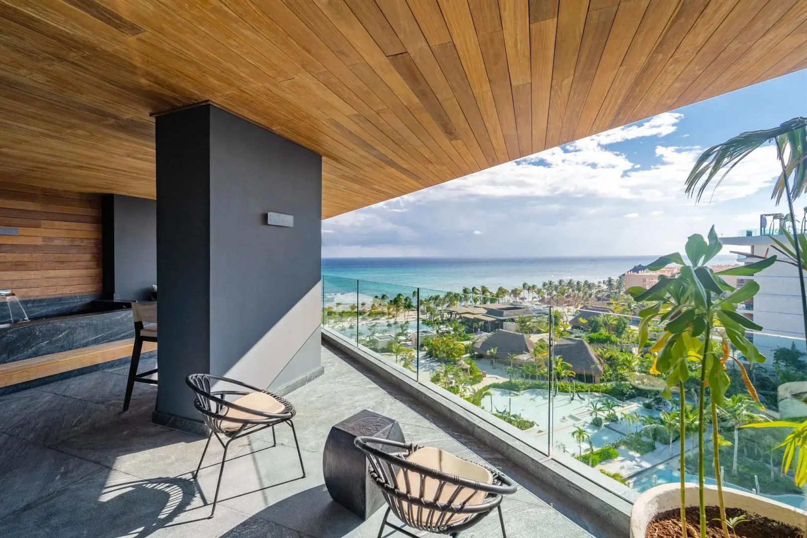 Blick von einer überdachten Terrasse mit Sitzstühlen auf die weitläufige Poollandschaft und das Meer.