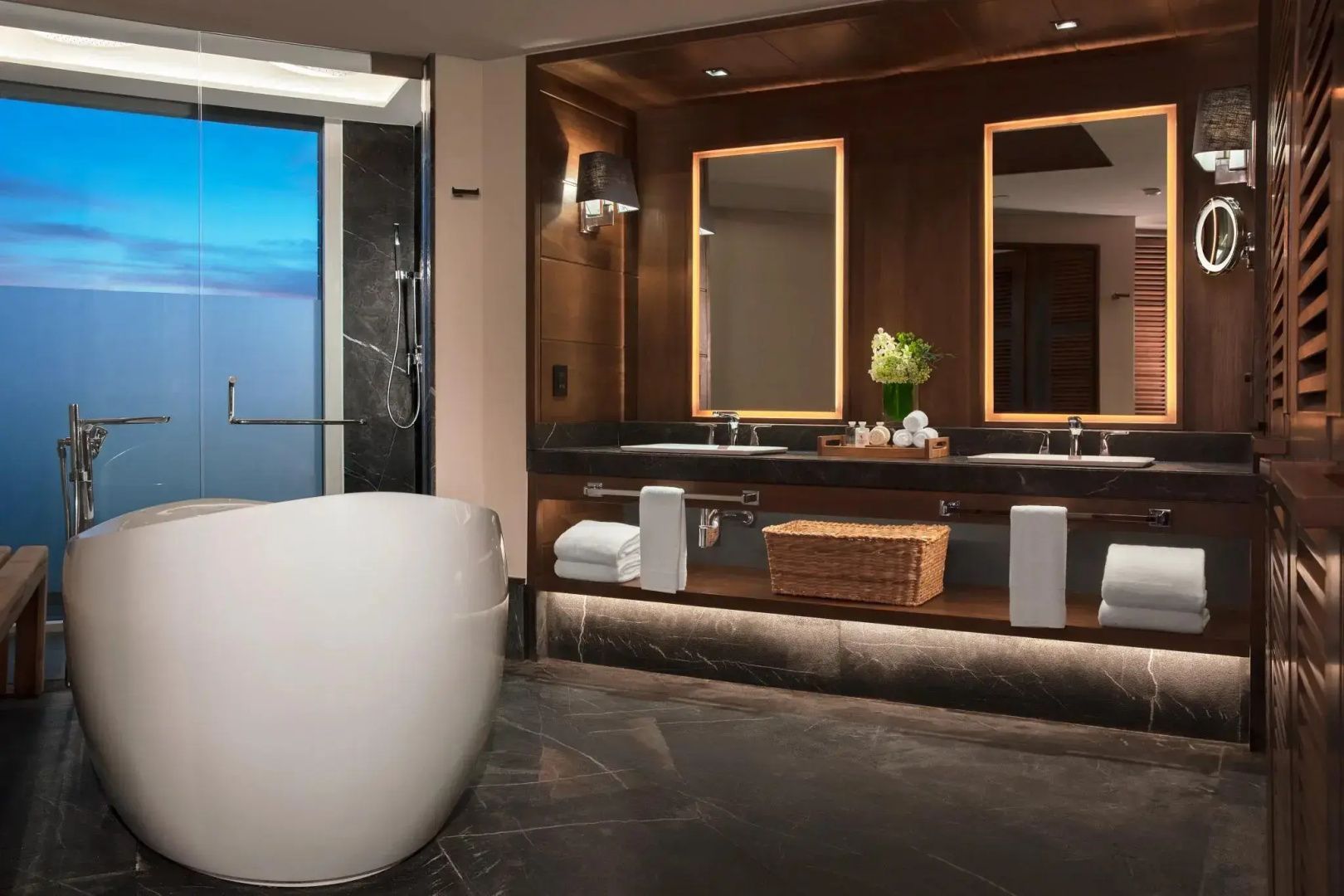 Badezimmer mit frei stehender Badewanne und Doppelwaschbecken vor beleuchteten Spiegeln.