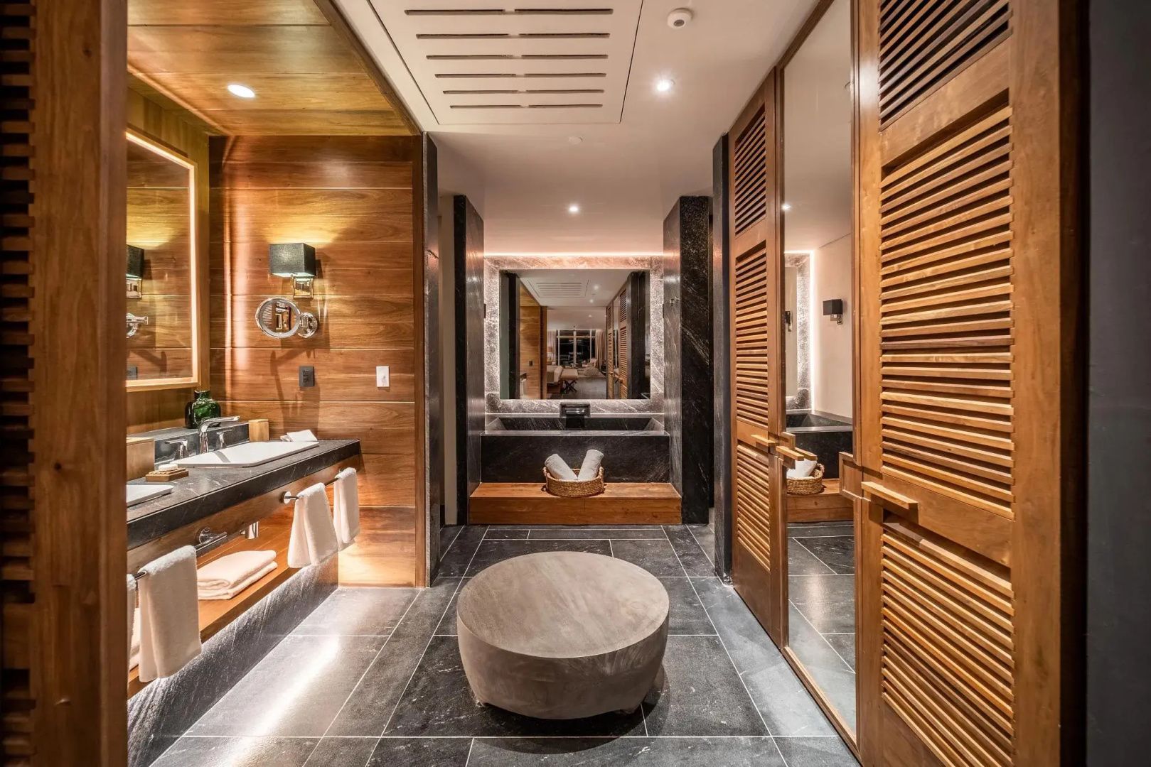 Badezimmerbereich mit rundem Hocker, Spiegel, Waschtisch und Schranktüren aus Holz.