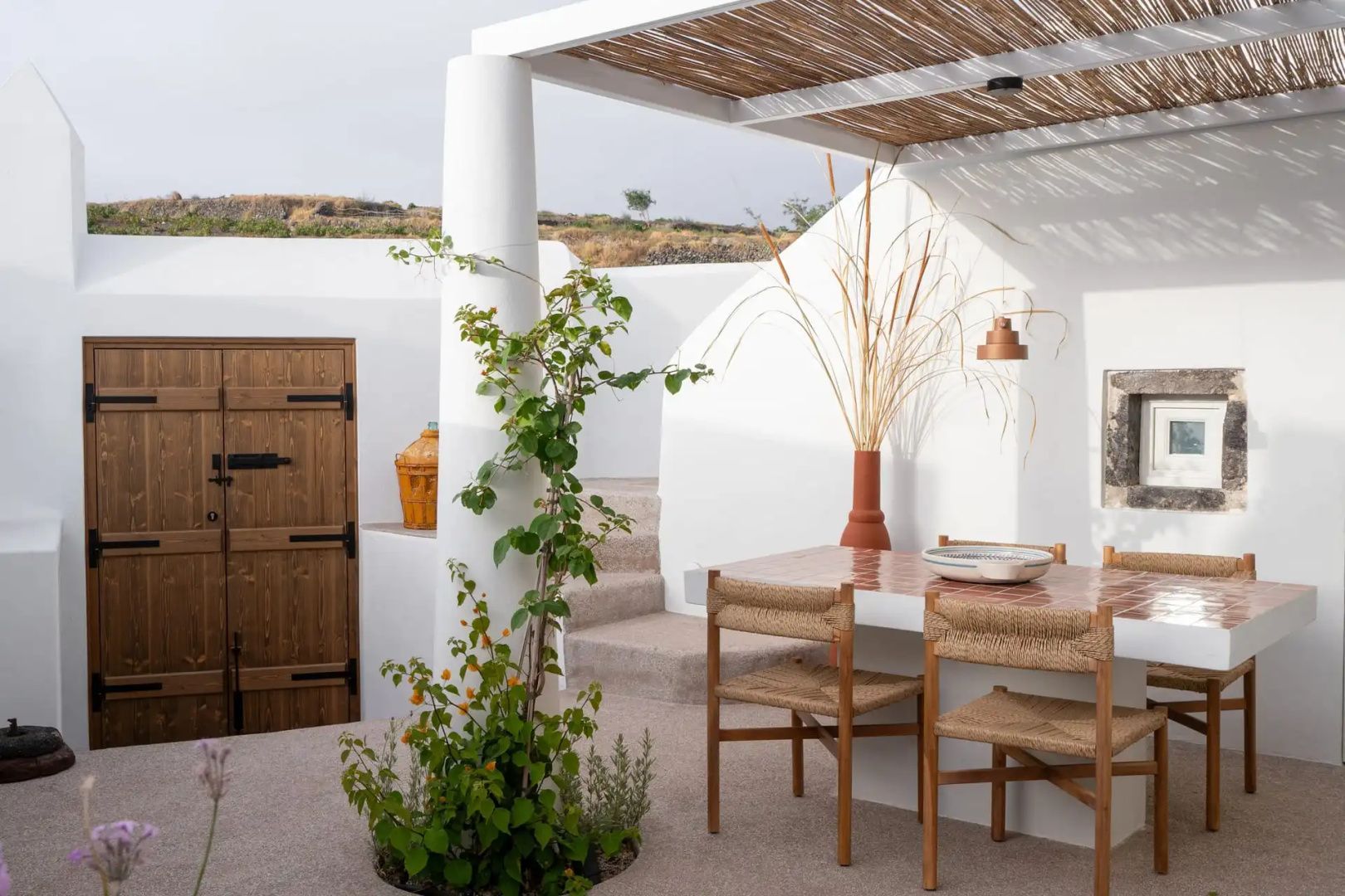 Holztisch mit Stühlen unter einer Pergola neben einer bepflanzten weißen Säule.