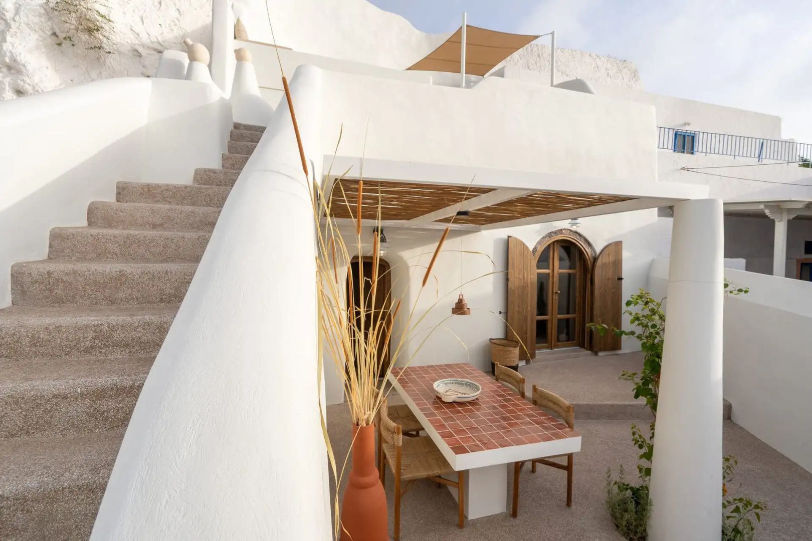 Weiße Gebäudefront mit Treppe zur oberen Terrasse und schattigem Esstisch.