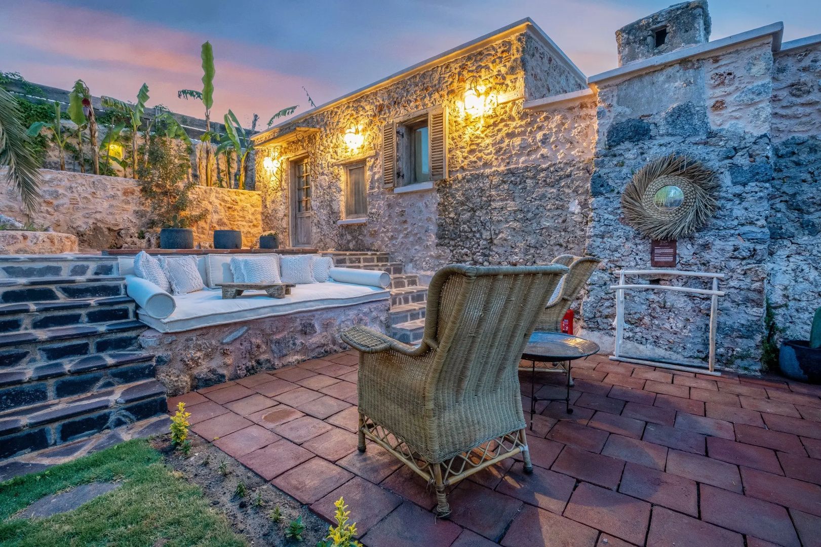 Gepflasterte Terrasse mit gemütlicher Loungeliege vor einer beleuchteten Natursteinwand.