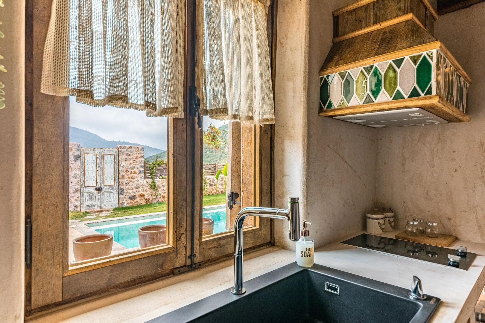 Blick durch ein Holzfenster mit Gardinen auf einen Pool und Steinmauern im Garten.