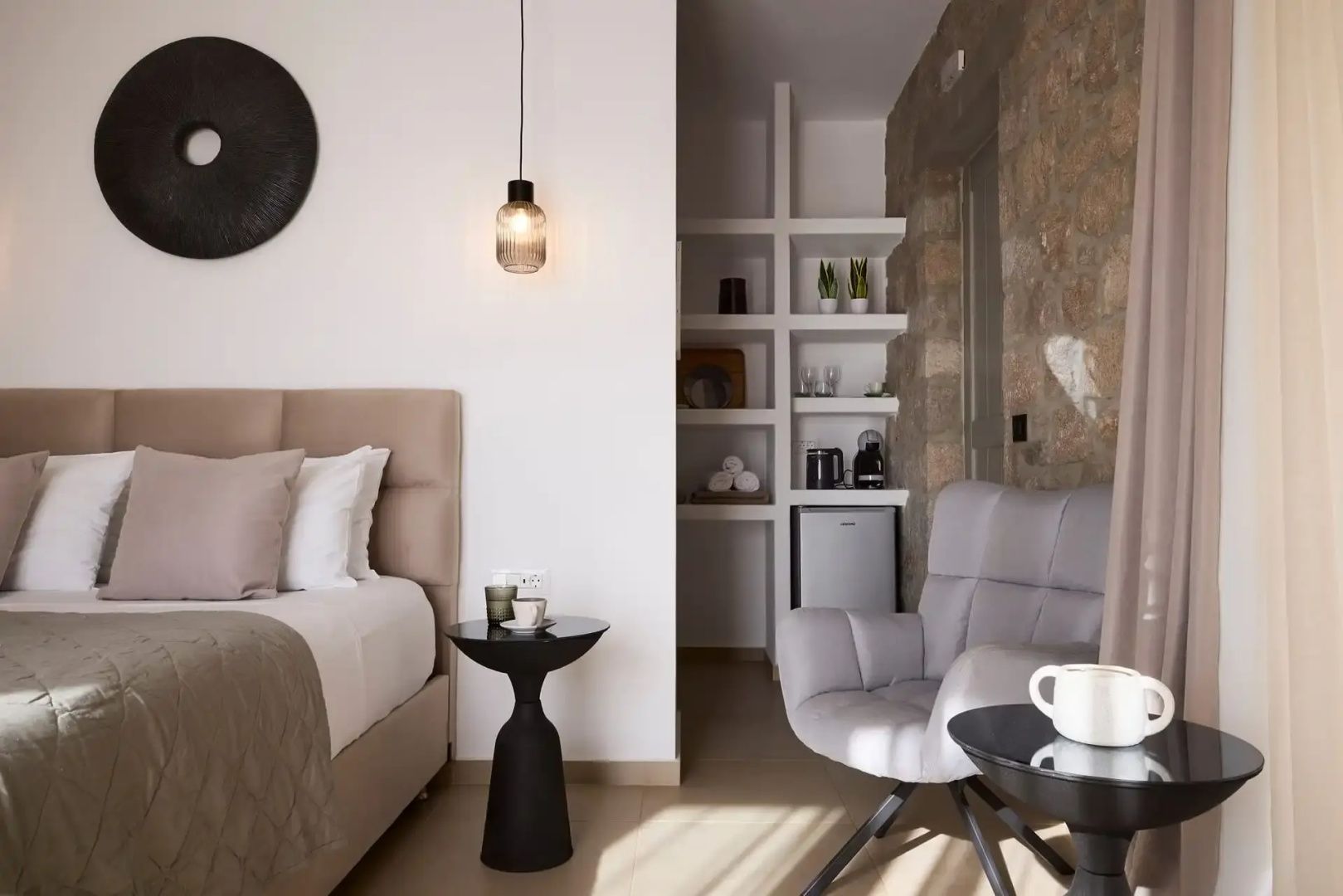 Modernes Hotelzimmer mit hellgrauem Sessel, Bett und offenem Regal vor Natursteinwand.