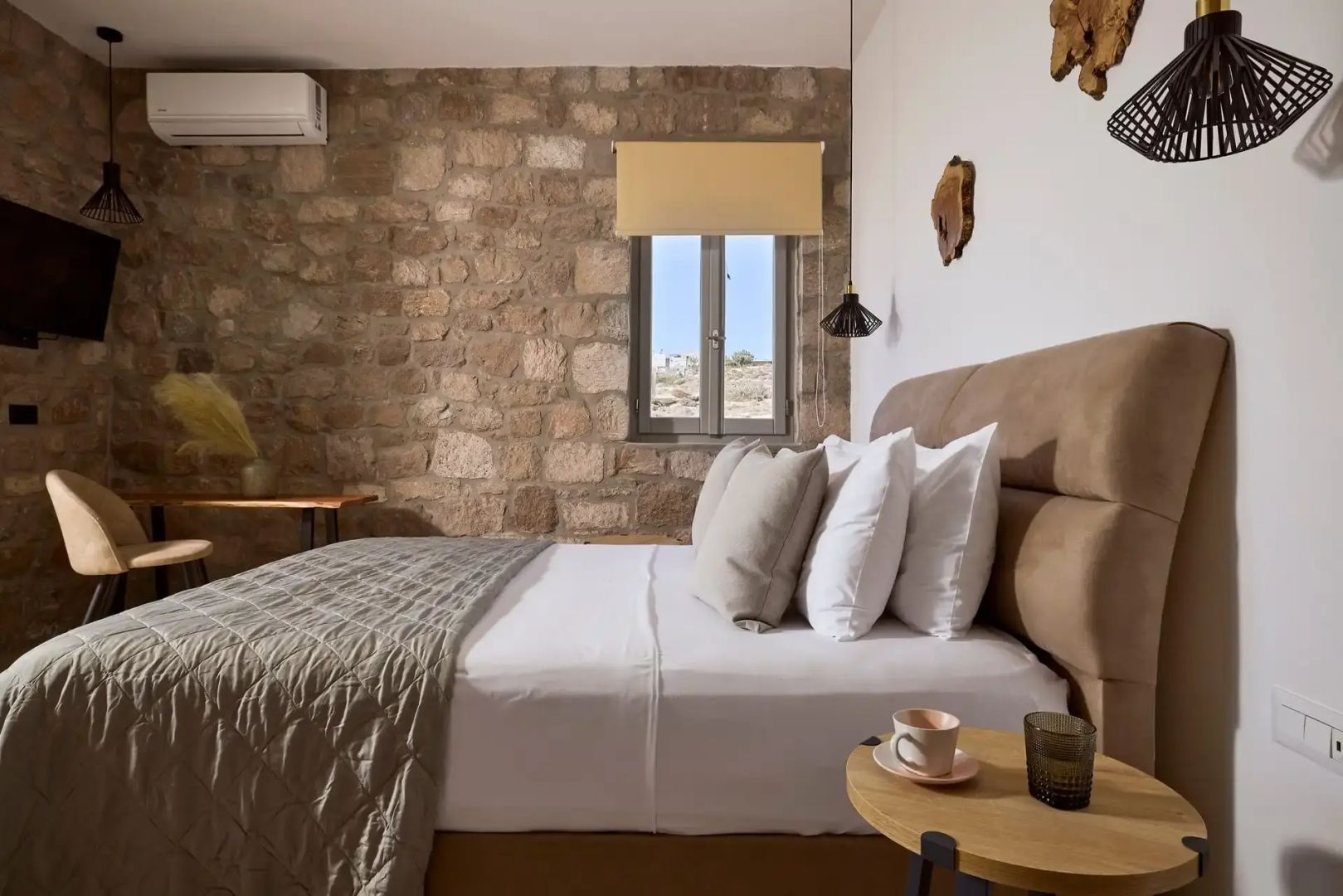Doppelbett mit Kissen, Natursteinwand, Beistelltisch und kleines Fenster im Hintergrund.