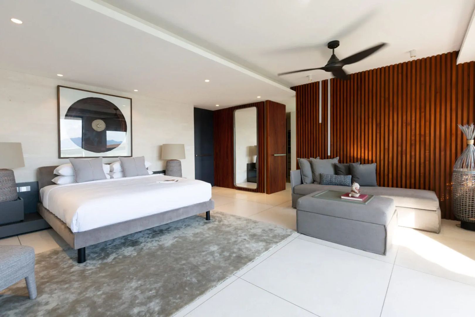 Schlafbereich einer Villa mit Doppelbett, grauem Sofa, Teppich und Holzverkleidung an der Wand.