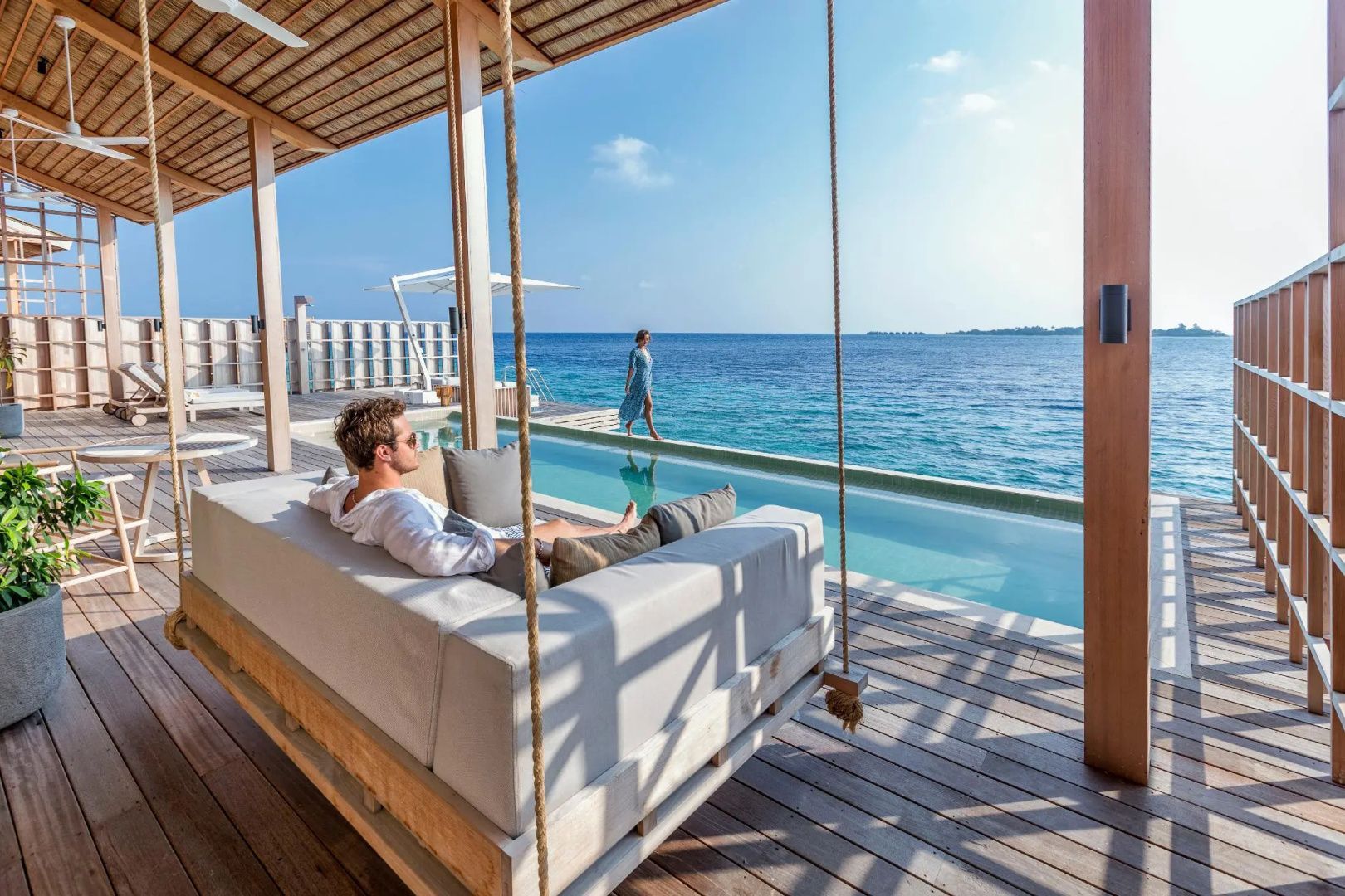 Personen auf einer Holzterrasse mit Hängeliege und schmalem Infinity Pool direkt am Meer.