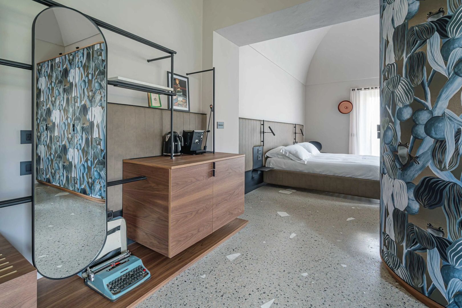 Schlafbereich einer Suite mit Holzkommode, Schreibmaschine, Terrazzoboden und großem Doppelbett.