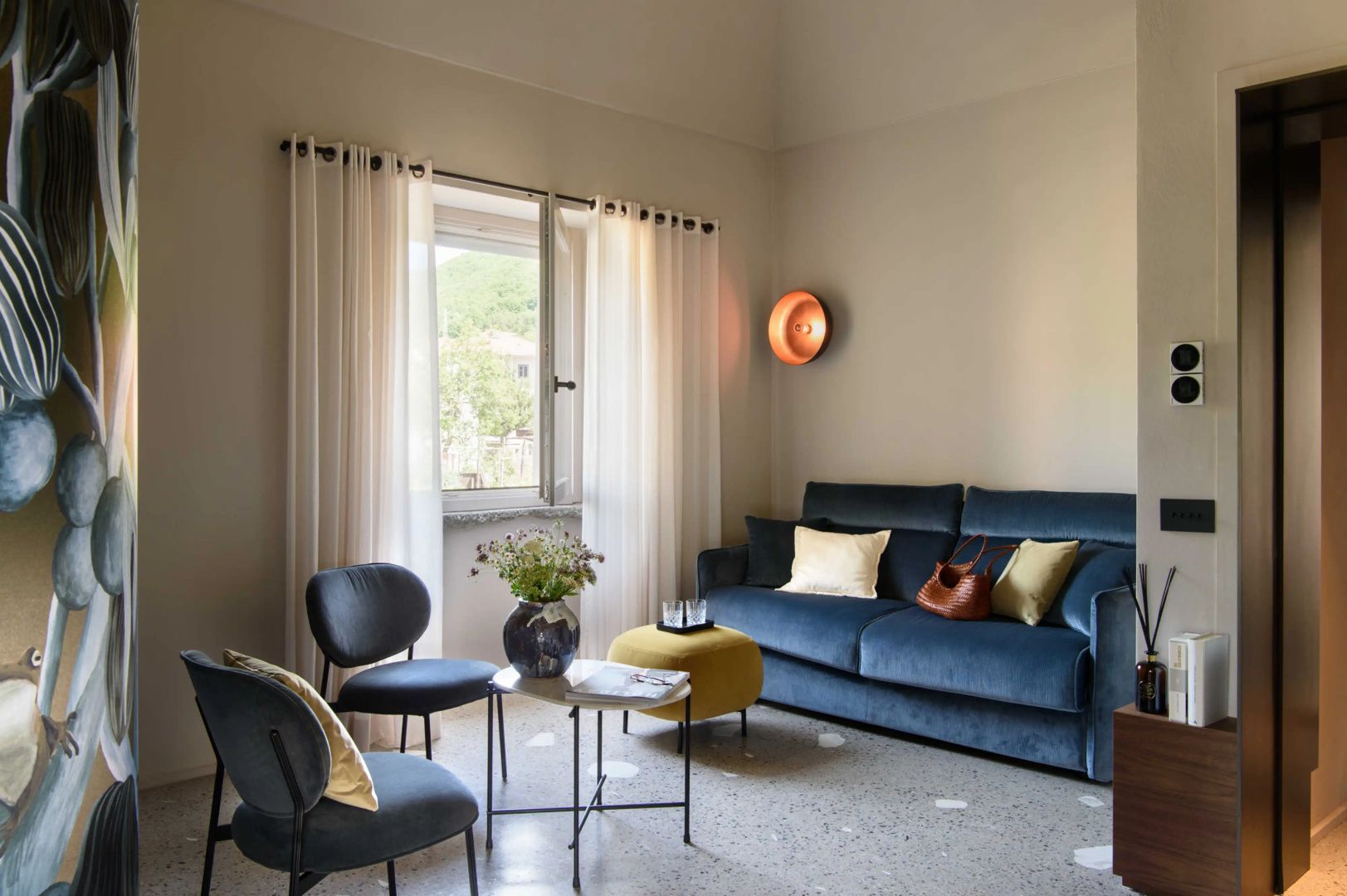 Wohnbereich mit blauem Samtsofa, gelbem Polsterhocker, zwei Sesseln und Fenster mit Vorhängen.