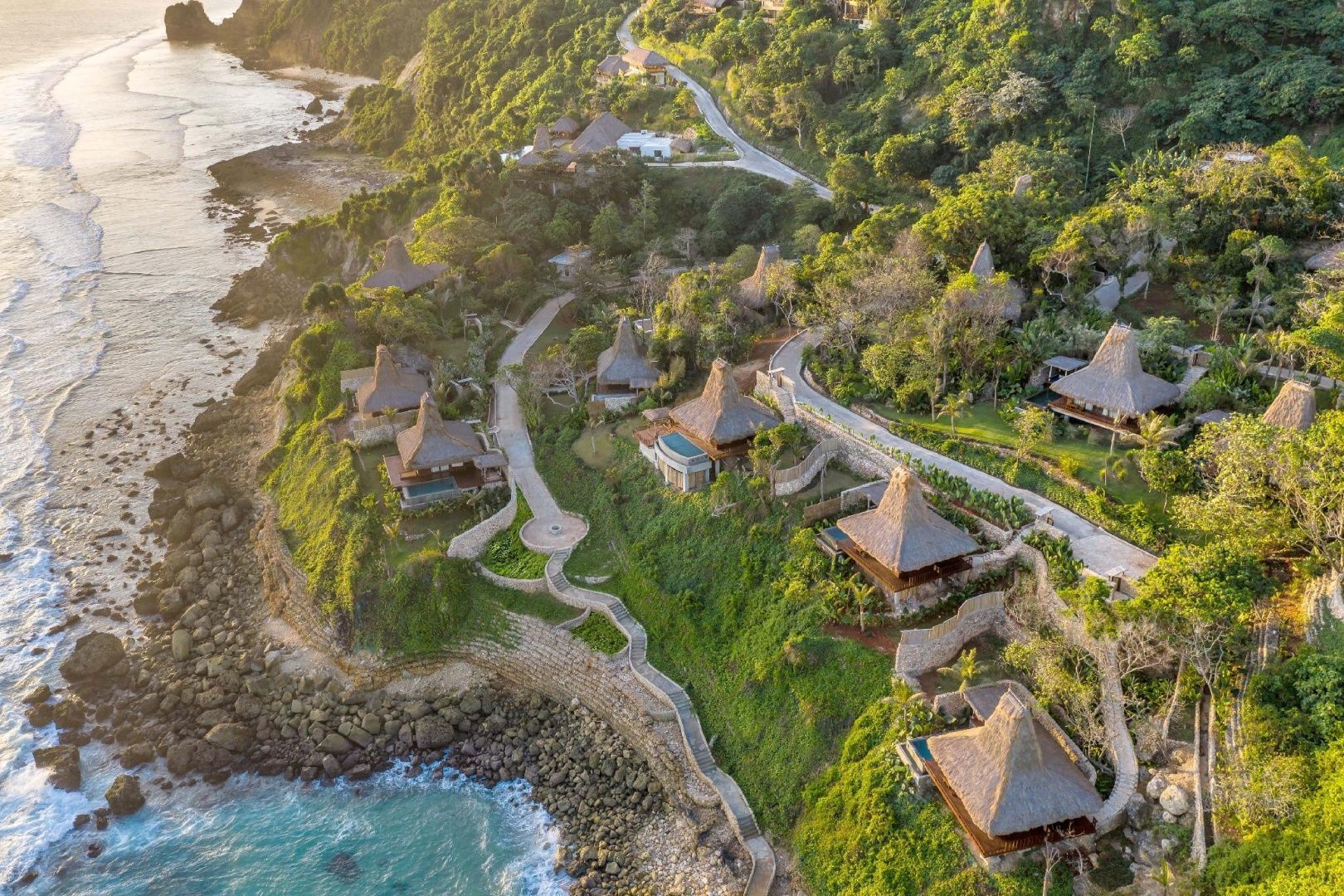 Luftaufnahme der Resortanlage mit Reetdach-Villas an einer Steilküste am Meer.