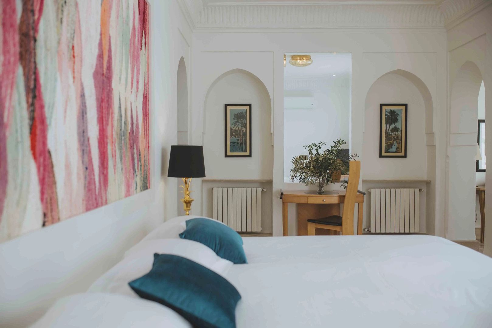 Schlafzimmer mit weiß bezogenem Bett, blauen Kissen, buntem Kunstwerk an der Wand und Wandnischen.