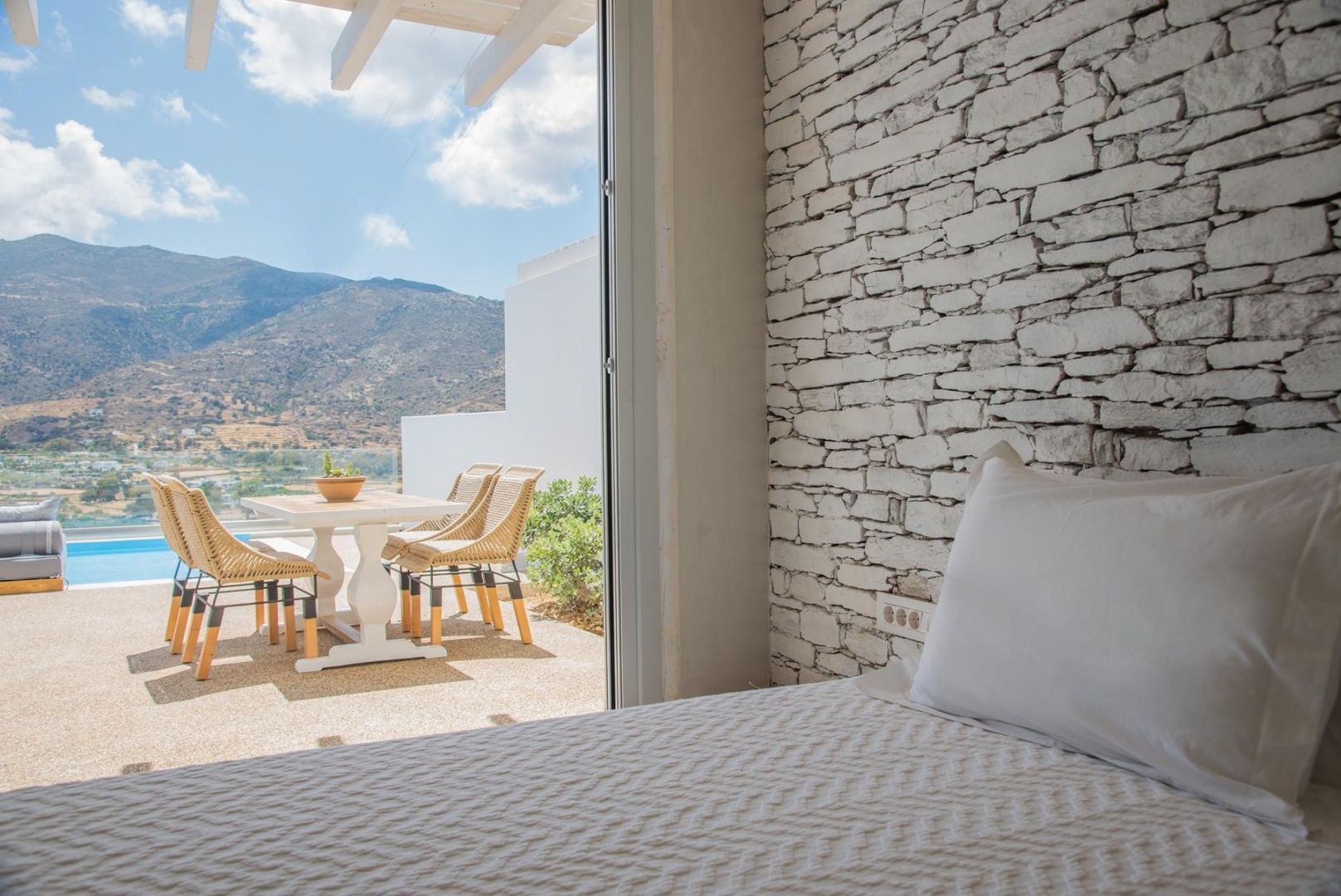 Blick vom Bett über die Terrasse mit Tisch und Stühlen auf die hügelige Landschaft.