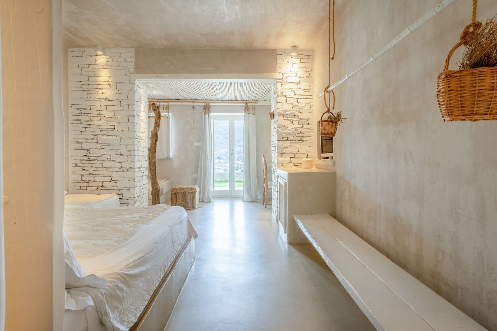 Helles Hotelzimmer mit Steinwänden, weißem Bett, Sitzbank und Balkontür im Hintergrund.