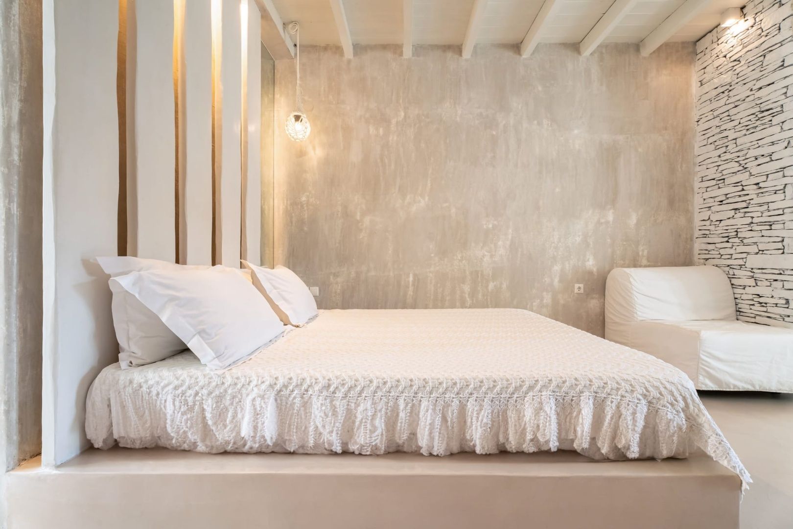 Doppelbett mit weißer Tagesdecke vor hellem Hintergrund und Holzdecke.