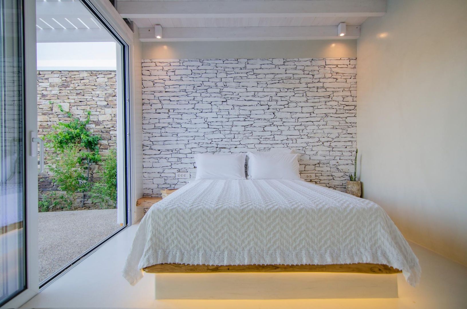 Modernes Hotelbett mit beleuchtetem Holzsockel vor einer Natursteinwand.