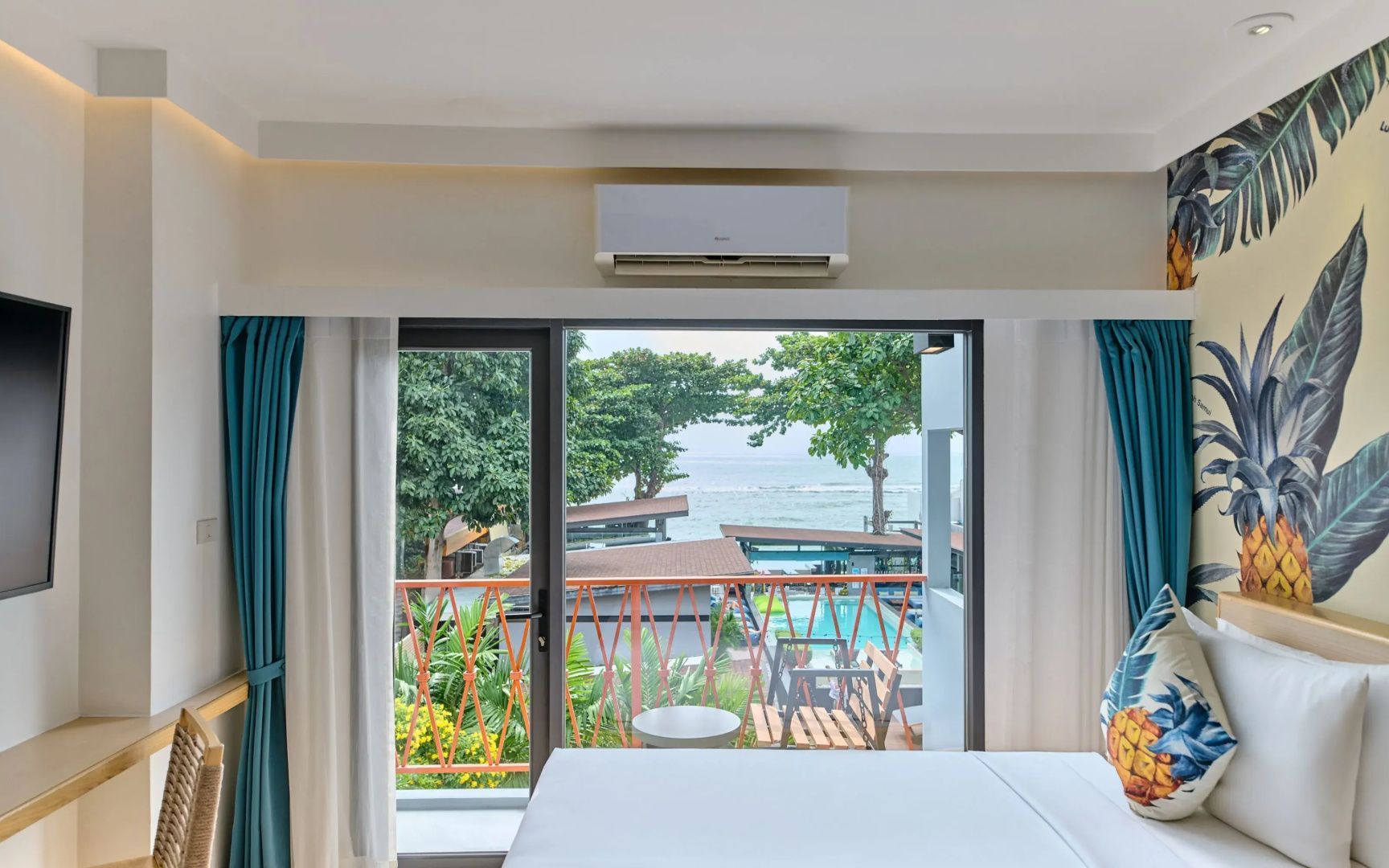 Blick durch eine Balkontür im Hotelzimmer auf grüne Bäume, den Swimmingpool und das Meer.