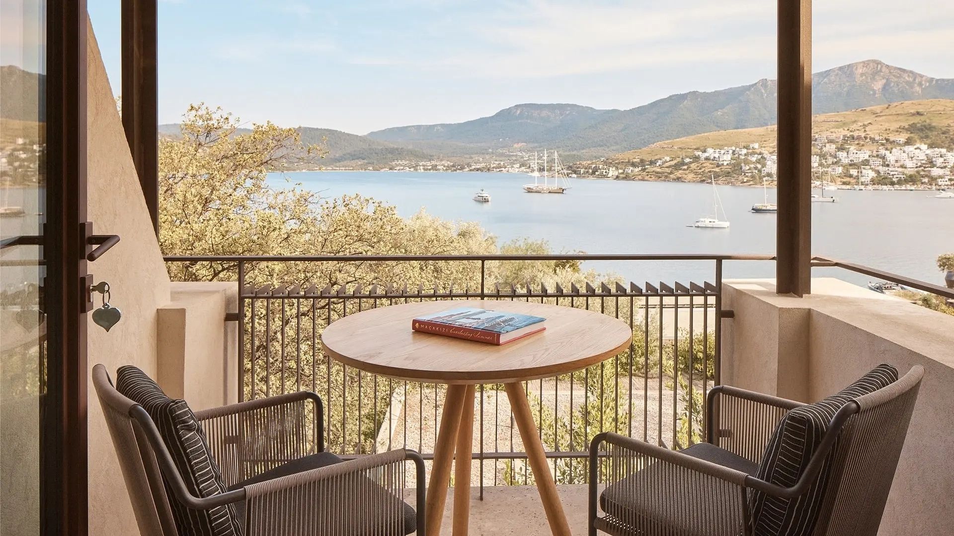 Balkon mit rundem Holztisch und zwei Stühlen mit Blick auf das Meer und Hügel.