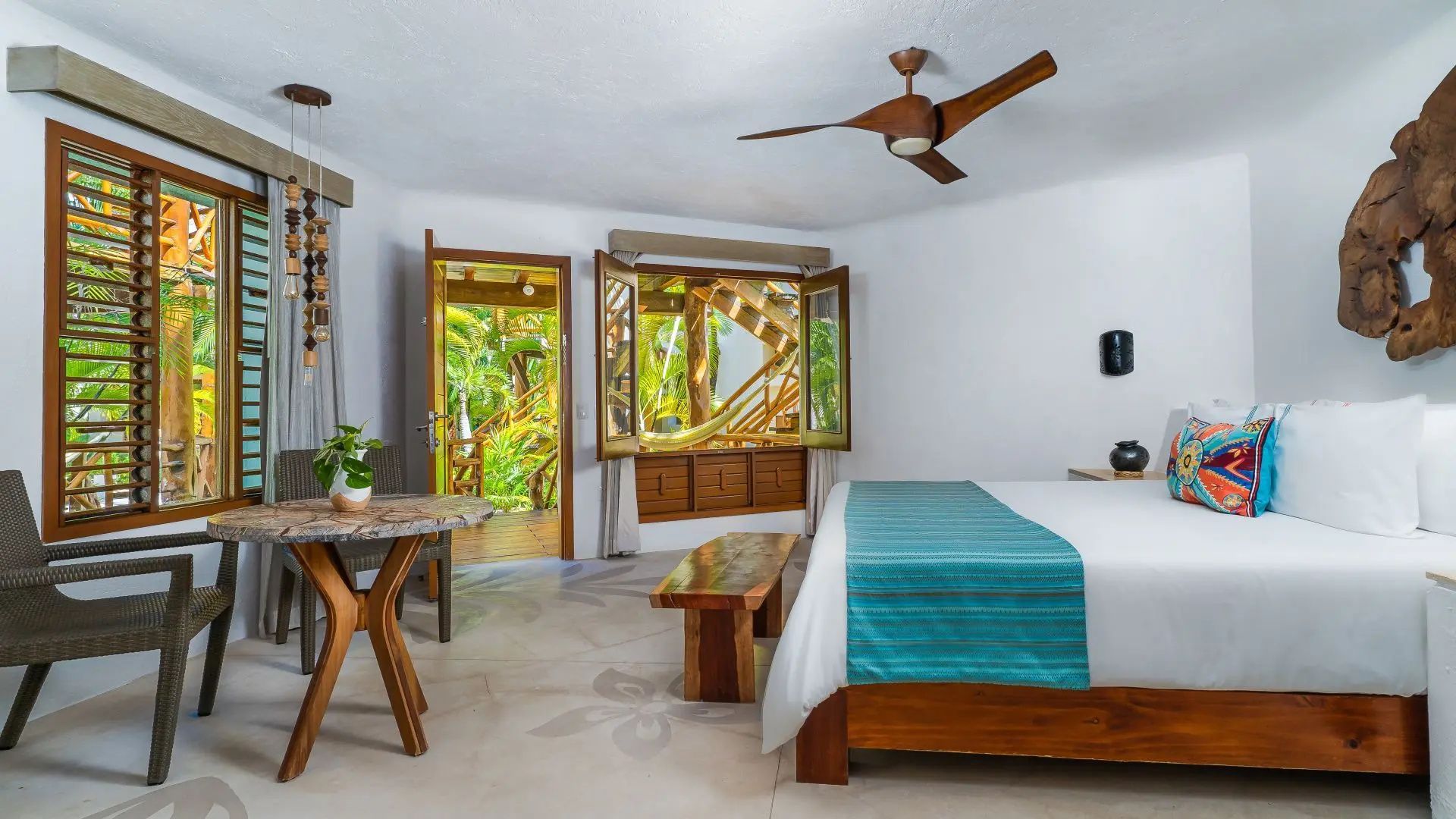 Helles Hotelzimmer mit Doppelbett, Ventilator und geöffneter Tür zum grünen Außenbereich.