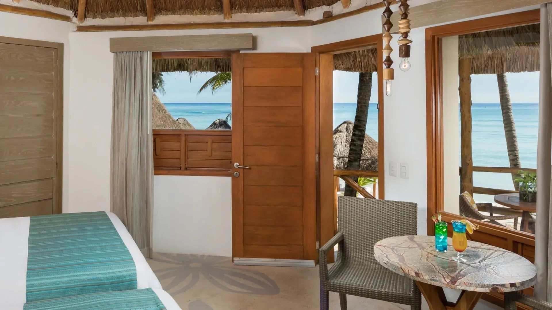 Blick durch ein geöffnetes Fenster und eine Tür auf das türkisblaue Meer und Palmen.