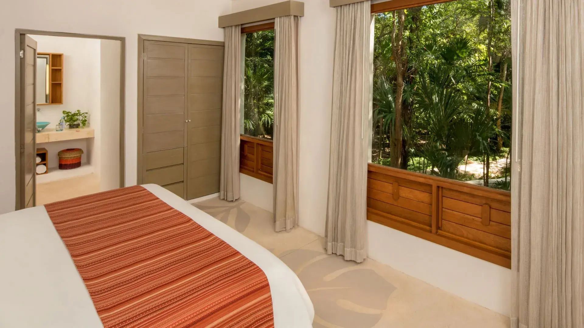 Schlafzimmer mit buntem Bettbezug, Holzschrank und Blick auf grüne Bäume.