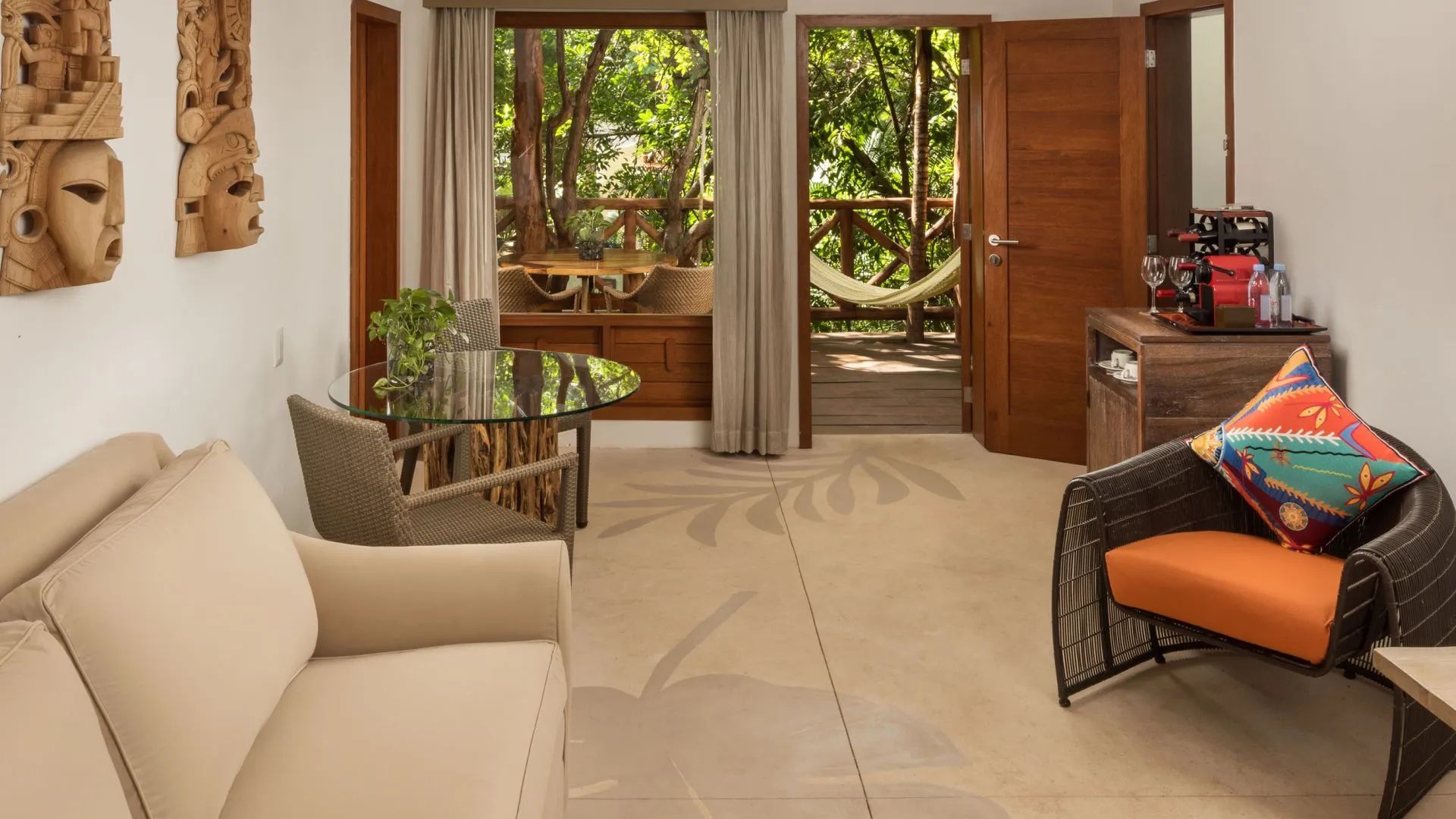 Wohnbereich eines Hotelzimmers mit Glastisch, Sesseln und Ausgang zur Holzterrasse mit Hängematte.