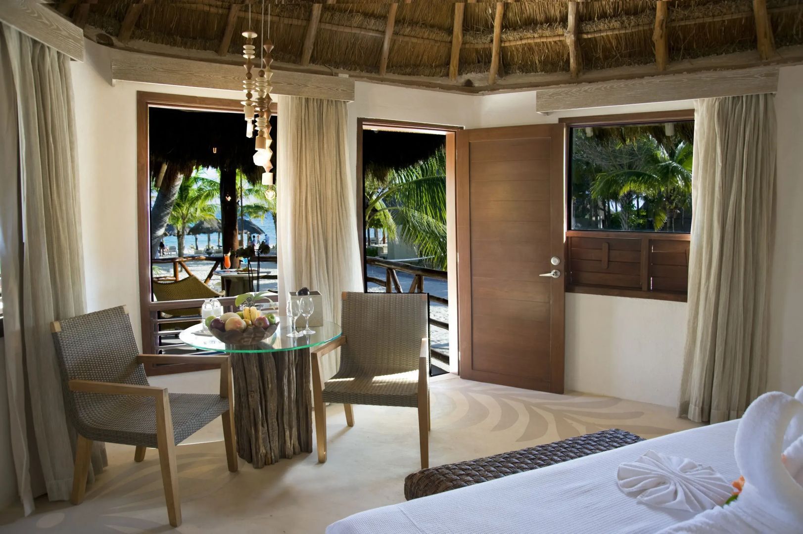 Bungalowzimmer mit Reetdach, Glastisch, Stühlen und Ausblick auf Palmen im Außenbereich.