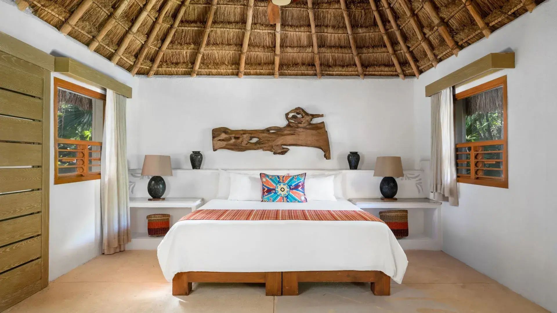 Doppelbett mit Reetdachdecke, bunten Kissen und einer Holzdekoration an der Wand.