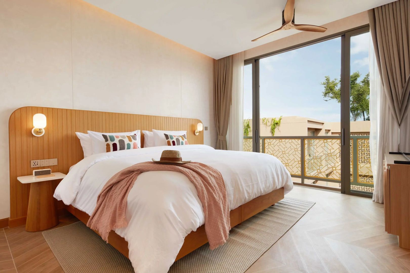 Helles Hotelzimmer mit Doppelbett, rosa Decke, Strohhut und großem Fenster zum Balkon.