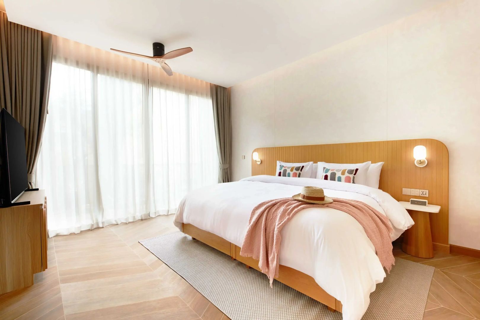 Doppelbett mit weißer Bettwäsche, rosa Decke und Strohhut vor großen Fenstervorhängen.