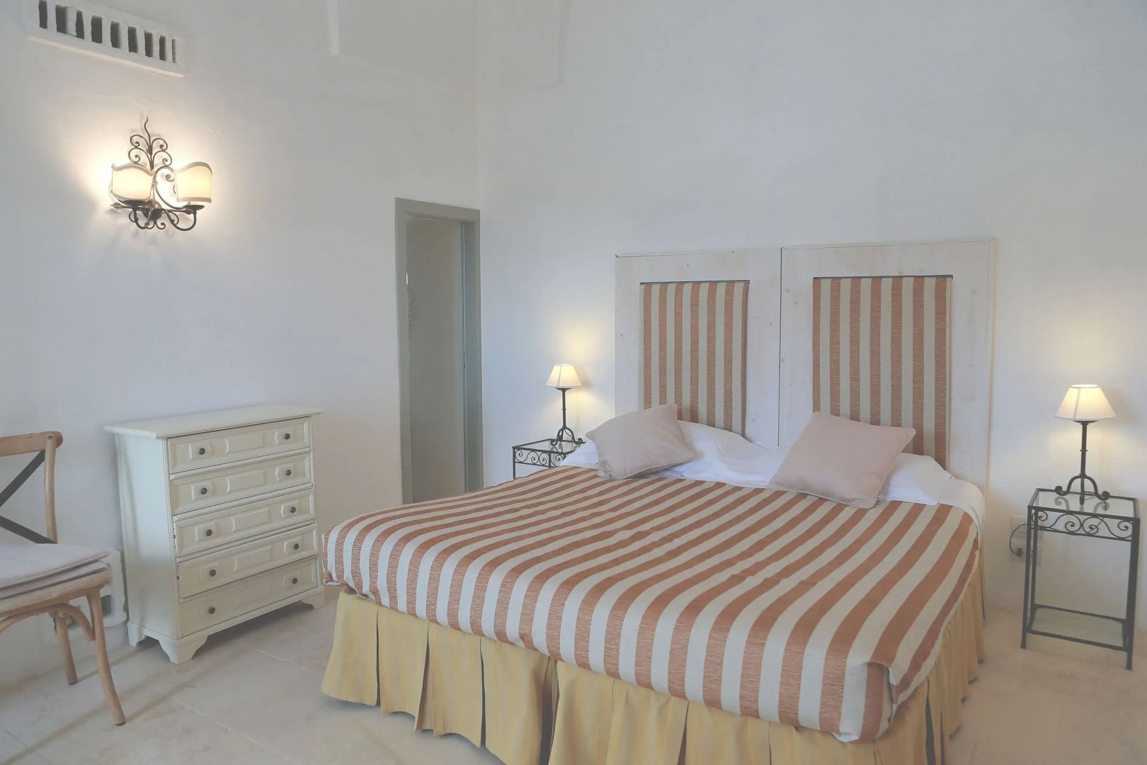 Schlafzimmer mit gestreiftem Doppelbett, beiger Kommode und Wandleuchte.