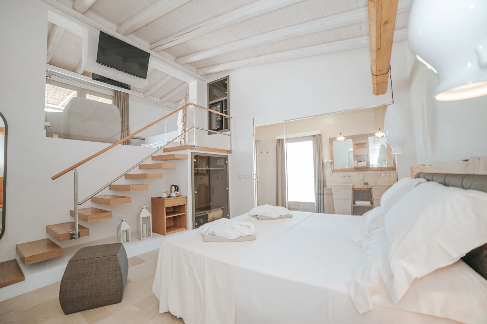 Helles zweigeschossiges Hotelzimmer mit Doppelbett, Holztreppe und offenem Badezimmerbereich.